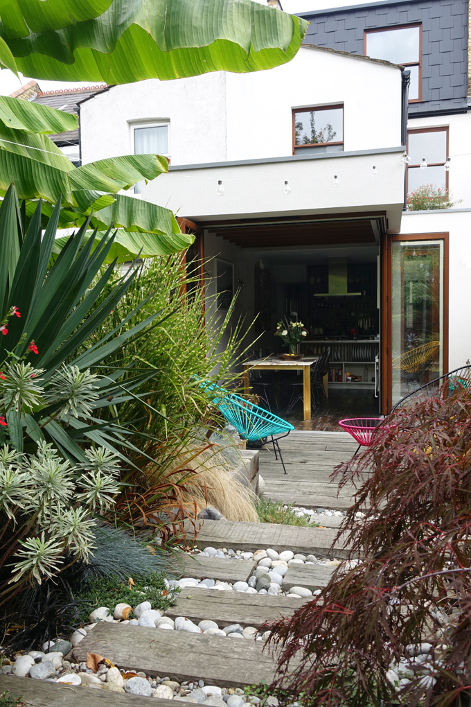 Rear ground floor extension with floor to ceiling bifolding doors, exposed beams and level threshold detail. Timber sleepers used for external landscaping, tropical planted garden and Acapulco chairs and table. Rooftop extension with basketweave tiling